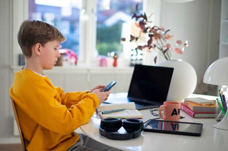 Interiørfoto av gutt som ser på smarttelefon foran PC og nettbrett
